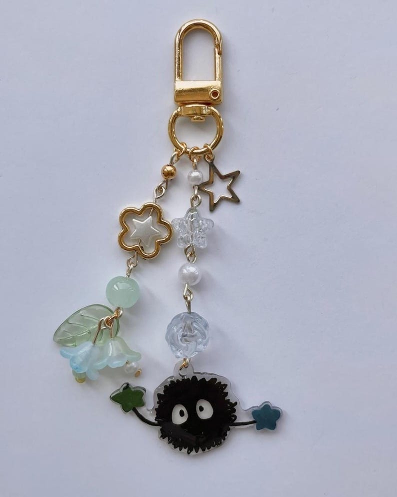 May include: A gold-colored keychain with various charms. Charms include a gold flower, a clear star, a white pearl, a blue flower, and a black soot sprite with white eyes. The keychain is attached to a gold clasp.
