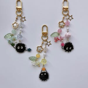 May include: Three gold-colored keychains with decorative charms. Each keychain features a clasp, star and flower charms, beads, and a black, round character with white eyes. The charms are in various colors, including pink, green, and yellow.