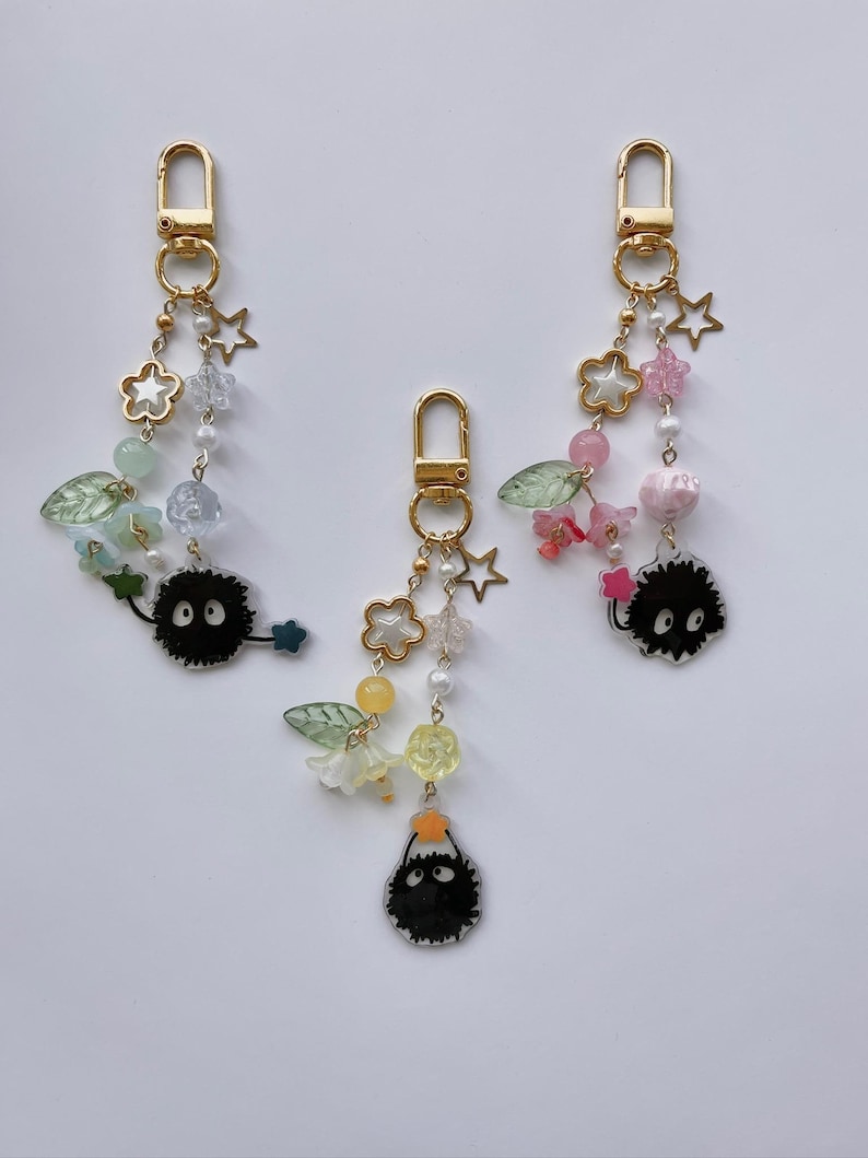 May include: Three gold-colored keychains with decorative charms. Each keychain features a clasp, star and flower charms, beads, and a black, round character with white eyes. The charms are in various colors, including pink, green, and yellow.
