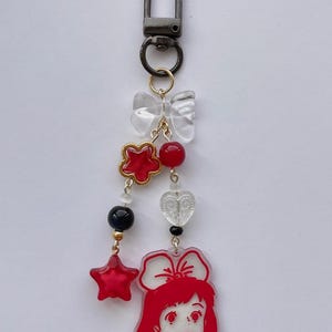 May include: A decorative keychain with a black clasp and a series of charms. The charms include a clear bow, red star and flower shapes, red and black beads, and a red-haired girl with a bow. The keychain is suspended on a gold chain.