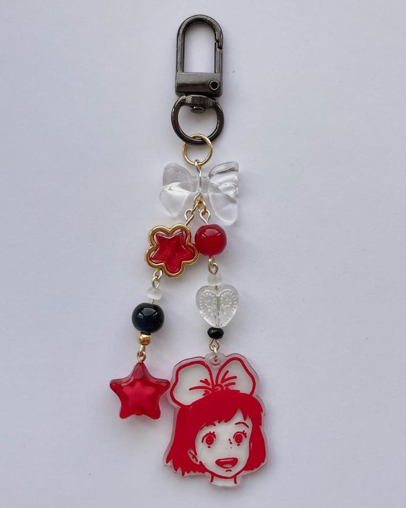 May include: A decorative keychain with a black clasp and a series of charms. The charms include a clear bow, red star and flower shapes, red and black beads, and a red-haired girl with a bow. The keychain is suspended on a gold chain.