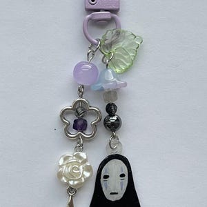 May include: A decorative keychain with a lavender clasp. The charm features a variety of beads, including purple, blue, and black, along with a flower, a star, a leaf, and a black figure with a white face.