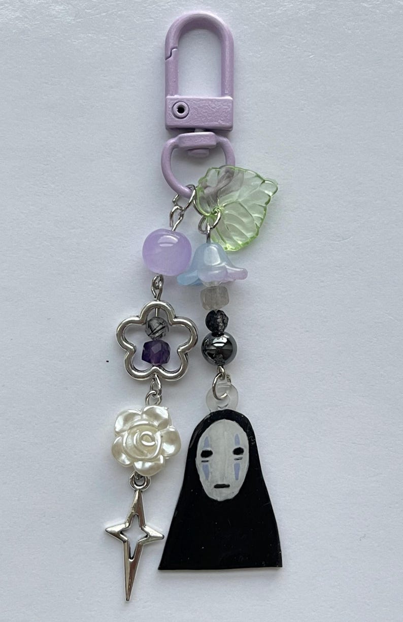 May include: A decorative keychain with a lavender clasp. The charm features a variety of beads, including purple, blue, and black, along with a flower, a star, a leaf, and a black figure with a white face.