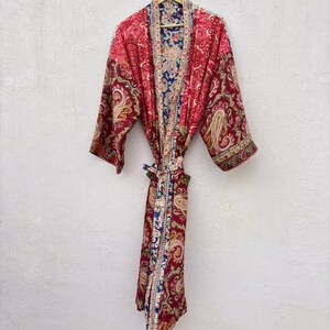 May include: A long-sleeved robe featuring a vibrant red and pink paisley pattern with contrasting blue and white floral trim along the front opening and collar. The garment includes a matching fabric tie belt at the waist and wide sleeves.