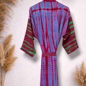May include: A tie-dye robe in shades of blue, red, and green hangs on a yellow hanger. The robe features a vertical striped pattern and a matching belt. The sleeves have a contrasting color pattern.