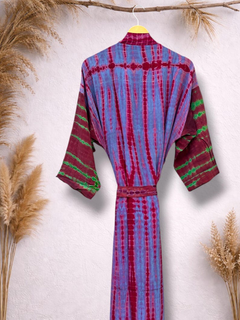 May include: A tie-dye robe in shades of blue, red, and green hangs on a yellow hanger. The robe features a vertical striped pattern and a matching belt. The sleeves have a contrasting color pattern.