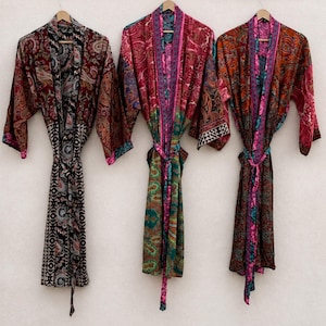 May include: Three long-sleeved robes hanging on wooden hangers, each featuring a distinct, intricate multi-colored paisley and geometric pattern. The robes are tied at the waist with matching fabric belts and display vibrant hues of red, black, green, and pink.