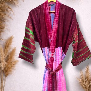 May include: A tie-dye robe in shades of burgundy, pink, and blue hangs on a yellow hanger. The robe features a wrap-around design with long sleeves and a matching belt. Green stripes accent the sleeves.