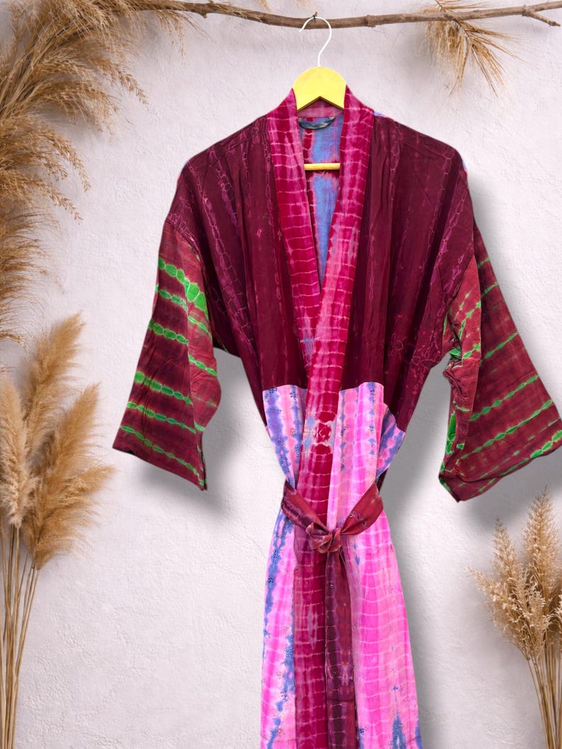 May include: A tie-dye robe in shades of burgundy, pink, and blue hangs on a yellow hanger. The robe features a wrap-around design with long sleeves and a matching belt. Green stripes accent the sleeves.