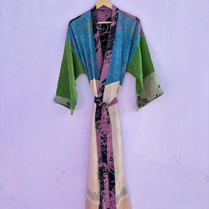 May include: A colorful, patchwork-style robe with long sleeves and a matching belt. The robe features panels of blue, green, purple, and cream fabrics with floral and geometric patterns. The robe is hanging on a wooden hanger against a light purple wall.