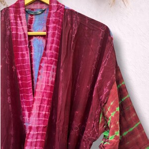 May include: A tie-dye robe in shades of burgundy, pink, and green hangs on a wooden hanger. The robe features a V-neck and long sleeves. The fabric has a soft, flowing appearance, with a mix of vertical and horizontal patterns.