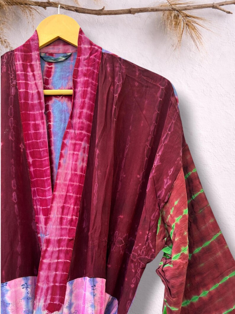 May include: A tie-dye robe in shades of burgundy, pink, and green hangs on a wooden hanger. The robe features a V-neck and long sleeves. The fabric has a soft, flowing appearance, with a mix of vertical and horizontal patterns.