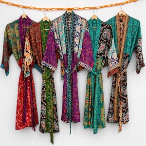 May include: Five long-sleeved robes hanging on wooden hangers from an orange chain. Each robe features a distinct patchwork design with vibrant paisley and floral patterns in shades of red, green, purple, teal, black, and gold with matching belts.