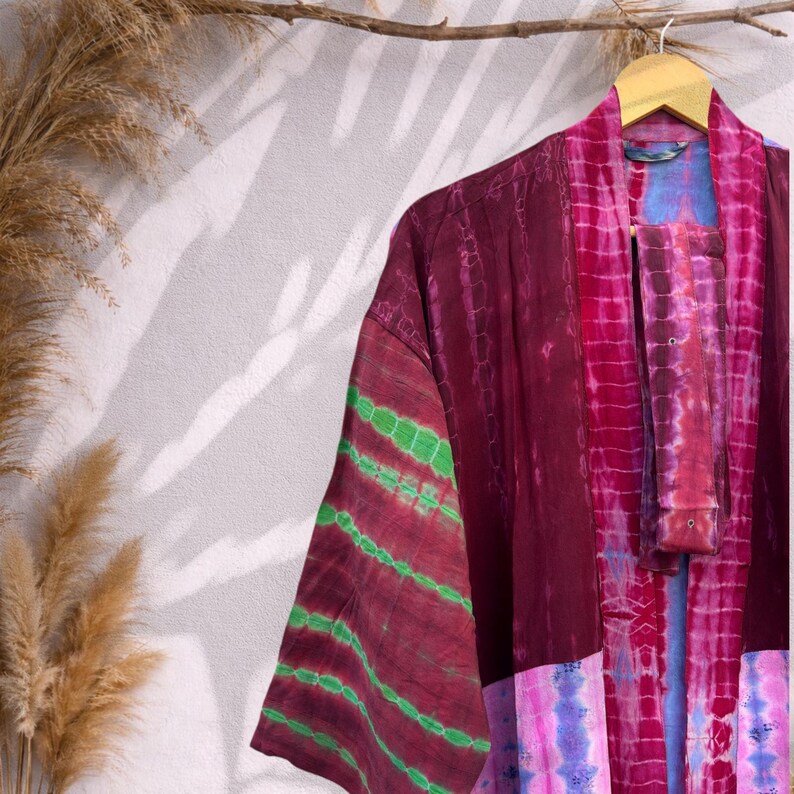 May include: A tie-dye kimono in shades of burgundy, pink, and green hangs on a wooden hanger. The kimono features a striped pattern on the sleeves and a contrasting panel at the bottom. The garment is made of a soft, flowing fabric.