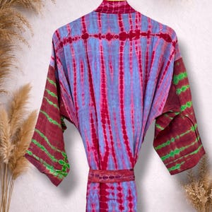 May include: A tie-dye robe in shades of blue, red, and maroon, hanging on a yellow hanger. The robe features vertical stripes and a matching belt. The sleeves have maroon and green accents. The robe is displayed against a neutral background.