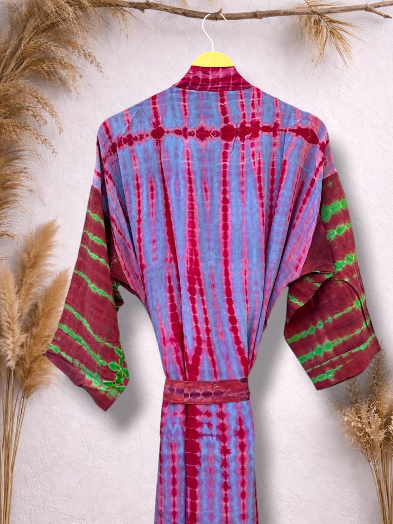 May include: A tie-dye robe in shades of blue, red, and maroon, hanging on a yellow hanger. The robe features vertical stripes and a matching belt. The sleeves have maroon and green accents. The robe is displayed against a neutral background.