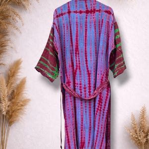 May include: A tie-dye robe in shades of blue and magenta hangs on a hanger. The robe has a matching belt and sleeves with green accents. The garment is displayed against a neutral background with dried plants.