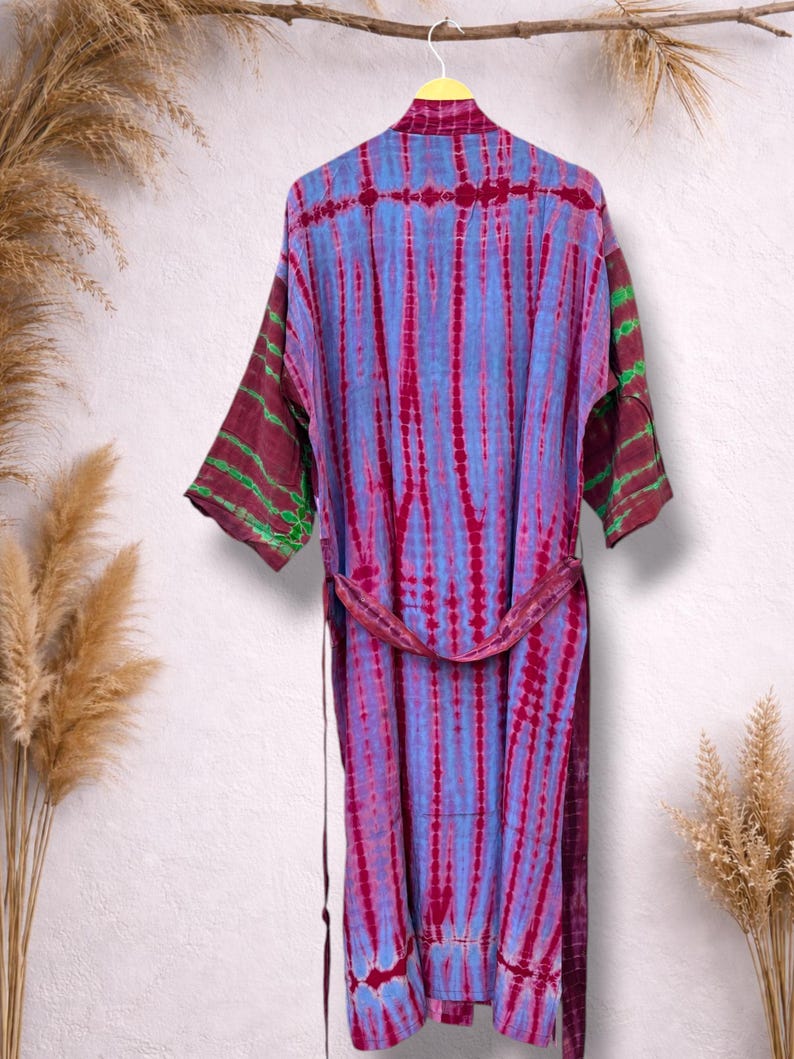 May include: A tie-dye robe in shades of blue and magenta hangs on a hanger. The robe has a matching belt and sleeves with green accents. The garment is displayed against a neutral background with dried plants.