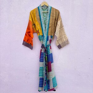 May include: A patchwork kimono robe in various colors, including orange, yellow, brown, and blue. The robe features a tie belt and is hanging on a wooden hanger against a light purple wall. The sleeves have different patterns and colors.
