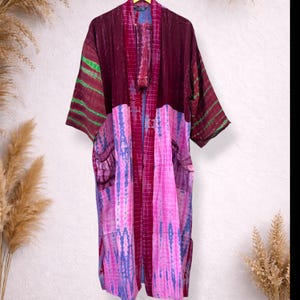 May include: A long, open-front kimono robe in shades of burgundy, pink, and blue. The robe features a tie-dye pattern on the lower half and striped accents on the sleeves. It has two pockets and a matching sash.