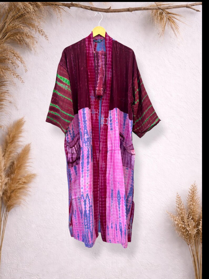 May include: A long, open-front kimono robe in shades of burgundy, pink, and blue. The robe features a tie-dye pattern on the lower half and striped accents on the sleeves. It has two pockets and a matching sash.