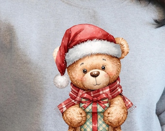 May include: A light grey sweatshirt with a festive teddy bear design. The bear is wearing a red Santa hat, a tartan scarf, and holding a wrapped gift. The design has a charming Christmas theme.
