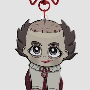 Pode incluir: Um chaveiro em estilo cartoon com um personagem com cabe&ccedil;a costurada, cabelo escuro e um la&ccedil;o vermelho. O personagem usa uma camisa branca com um la&ccedil;o vermelho e uma saia xadrez. O chaveiro tem um fecho em forma de la&ccedil;o vermelho.