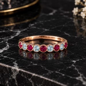 May include: A rose gold band ring featuring an alternating pattern of round-cut red rubies and light blue aquamarine gemstones. Each stone is set in a milgrain-detailed bezel setting, creating a continuous row of colorful gems along the band.