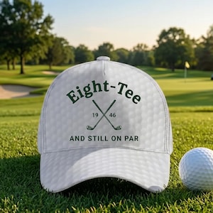 K&ouml;nnte beinhalten: Wei&szlig;e Golfkappe mit dem Text "Eight-Tee 1946 AND STILL ON PAR" in Gr&uuml;n. Die Kappe hat eine Golfballstruktur und gekreuzte Golfschl&auml;ger. Ein wei&szlig;er Golfball liegt auf dem gr&uuml;nen Gras.