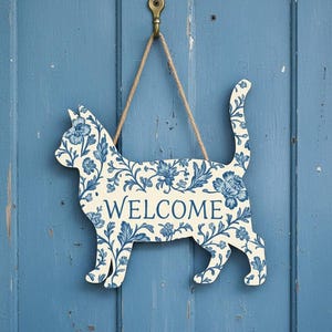 May include: A cat-shaped wall sign featuring a cream-colored base with a blue floral pattern and the word WELCOME printed in blue. The sign is suspended by a brown twine cord from a metal hook, displaying a decorative silhouette of a feline.