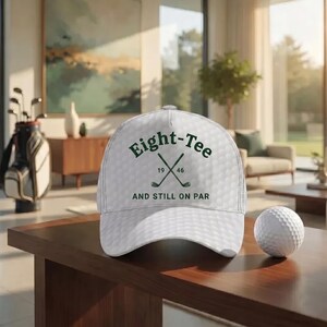 K&ouml;nnte beinhalten: Wei&szlig;e Golfkappe mit dem Text "Eight-Tee" in Dunkelgr&uuml;n, mit gekreuzten Golfschl&auml;gern und den Jahren "19" und "46". Die Kappe hat ein Golfballmuster. Ein wei&szlig;er Golfball liegt neben der Kappe auf einer Holzoberfl&auml;che.