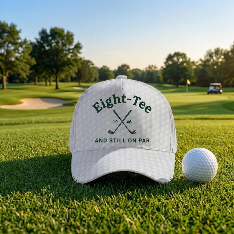 K&ouml;nnte beinhalten: Wei&szlig;e Golfkappe mit dem Text "Eight-Tee 1946 AND STILL ON PAR" in Gr&uuml;n. Die Kappe hat eine Golfballstruktur und gekreuzte Golfschl&auml;ger. Ein wei&szlig;er Golfball liegt auf dem gr&uuml;nen Gras.