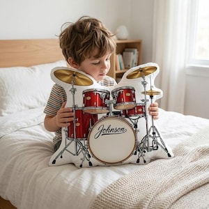 May include: A drum kit-shaped pillow with red drums, gold cymbals, and silver stands. The bass drum has the word "Johnson" printed on it. The pillow is on a white bed.