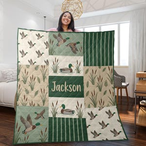 May include: A quilted blanket featuring a patchwork design with mallard ducks in flight and swimming among cattails. The colour palette includes shades of green, beige, and cream. The centre panel displays the name Jackson in white lettering.
