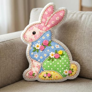Peut inclure: Un coussin décoratif en forme de lapin avec un motif patchwork. Les oreilles sont roses avec des motifs floraux blancs, et le corps présente des sections bleues, vertes et jaunes ornées de fleurs brodées et de perles. Le lapin a un œil en bouton noir.