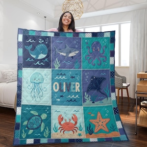 May include: A patchwork quilt with marine life illustrations. The squares show a whale, shark, octopus, jellyfish, stingray, turtle, crab, and starfish. The name "OLIVER" is in the centre. The quilt is in shades of blue and teal, with a quilted pattern.