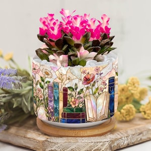 Puede incluir: Una maceta de cerámica decorativa con un diseño de vidriera que representa libros y flores. La maceta contiene una suculenta con flores de color rosa brillante. La maceta está sobre una base de madera, creando una exhibición encantadora.