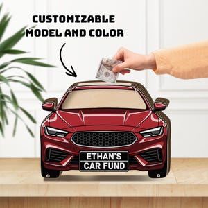 May include: A red car-shaped wooden money box with a clear front panel and a black license plate that reads ETHAN'S CAR FUND. The text CUSTOMIZABLE MODEL AND COLOR is printed above the car, which has a slot for inserting paper currency.