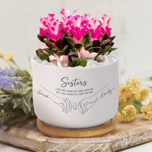 Könnte beinhalten: Ein weißer Keramiktopf mit einer leuchtend rosa blühenden Pflanze. Der Topf trägt das Wort "Sisters" und eine Botschaft sowie die Namen "Sarah" und "Emily". Der Topf steht auf einem Holzsockel.