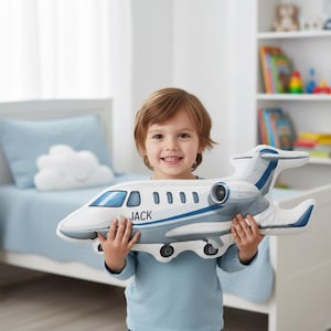 Peut inclure: Un coussin en forme d'avion blanc et gris avec des d&eacute;tails bleus et le nom "JACK" imprim&eacute; sur le c&ocirc;t&eacute;. Le coussin pr&eacute;sente un design d&eacute;taill&eacute;, avec des fen&ecirc;tres et des d&eacute;tails de r&eacute;acteur. Un enfant tient le coussin.