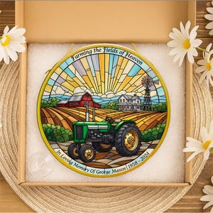 May include: A stained glass window featuring a green tractor in a farm scene with a red barn, farmhouse, and windmill. The text reads "Farming the Fields of Heaven" and "In Loving Memory of George Mason 1958-2024". The window is in a cardboard box.