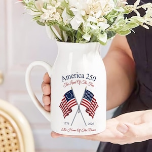 May include: White ceramic pitcher with a handle, featuring the text "America 250" and crossed American flags. The pitcher is filled with white flowers. The design includes the years 1776 and 2026.