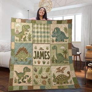 May include: A quilted blanket featuring a grid of sage green and beige squares with illustrations of various dinosaurs, including a T-Rex, Triceratops, and Stegosaurus, alongside a central square displaying the name JAMES in bold letters.