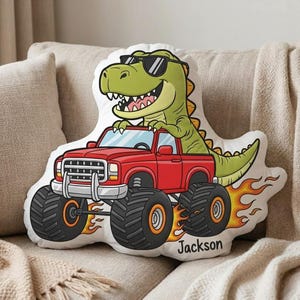 Pode incluir: Uma almofada branca com um dinossauro de desenho animado usando óculos de sol dirigindo um caminhão monstro vermelho. O caminhão tem pneus pretos grandes e chamas. O nome "Jackson" está impresso na almofada.