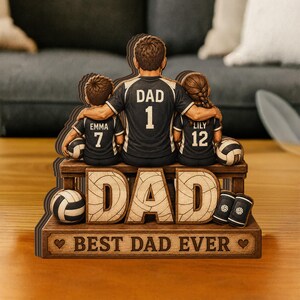 May include: A wooden tabletop display featuring a central figure wearing a jersey labeled DAD 1, with two children in jerseys labeled EMMA 7 and LILY 12. The base features the text DAD and BEST DAD EVER, accompanied by volleyballs and knee pads.