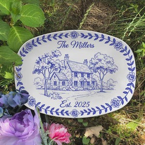 Pode incluir: Prato oval branco com uma borda floral azul e uma ilustração azul detalhada de uma casa com árvores. O texto "The Millers" e "Est. 2025" é exibido em azul. O prato é cercado por vegetação e flores.