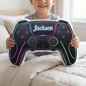 May include: A black and white video game controller-shaped pillow with the name "Jackson" in white script. The pillow has colorful neon accents and is held by a person.