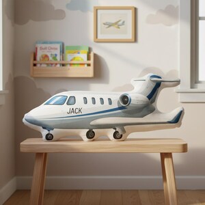 Peut inclure: Un coussin en forme d'avion blanc et gris avec des d&eacute;tails bleus et le nom "JACK" imprim&eacute; sur le c&ocirc;t&eacute;. Le coussin est pos&eacute; sur un banc en bois dans une chambre d'enfant, avec une image encadr&eacute;e d'un avion et une &eacute;tag&egrave;re en arri&egrave;re-plan.
