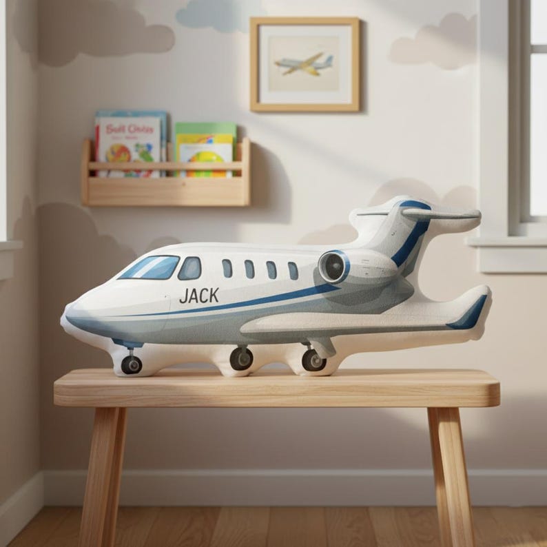 Peut inclure: Un coussin en forme d'avion blanc et gris avec des d&eacute;tails bleus et le nom "JACK" imprim&eacute; sur le c&ocirc;t&eacute;. Le coussin est pos&eacute; sur un banc en bois dans une chambre d'enfant, avec une image encadr&eacute;e d'un avion et une &eacute;tag&egrave;re en arri&egrave;re-plan.