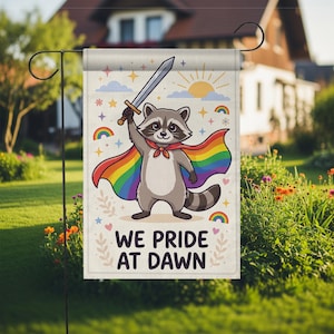 Puede incluir: Una bandera de jardín con un mapache de dibujos animados con una capa de arcoíris y una espada. La bandera tiene el texto "WE PRIDE AT DAWN" con arcoíris, estrellas y un sol. La bandera se muestra en un jardín.